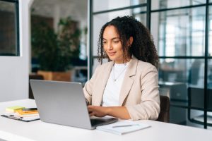 Woman uses laptop. Successful curly haired hispanic or brazilian woman in stylish elegant clothes, office employee, secretary or hr manager, using a laptop while sitting at her workplace, working